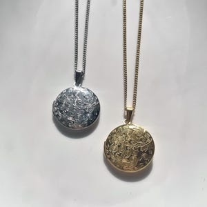 May include: Two round locket necklaces, one in silver tone and one in gold tone, each featuring an intricate engraved floral pattern on the front surface. Both lockets are suspended from matching thin metal link chains of the same colour.