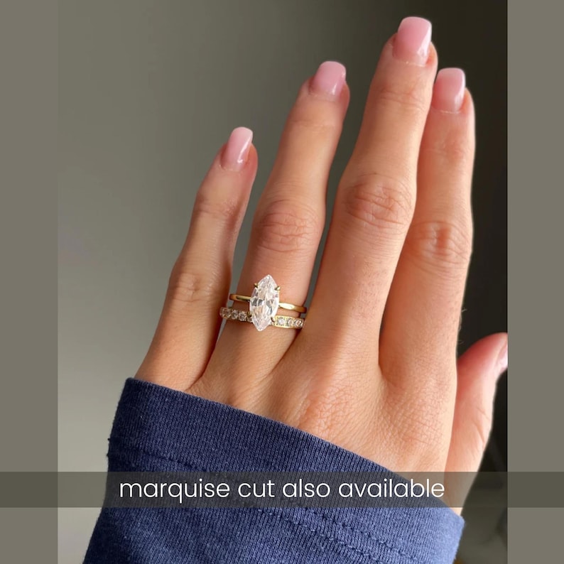 K&ouml;nnte beinhalten: Ein Goldring-Set bestehend aus einem Ring mit einem klaren Marquise-Schliff-Stein auf einer schlichten Schiene und einem zweiten Ring, der mit kleinen runden klaren Steinen besetzt ist. Der Text "marquise cut also available" steht unten.