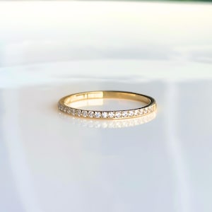 May include: A gold band ring with a row of small, clear gemstones set around the band. The ring is displayed on a white surface, reflecting light. The ring is a delicate piece of jewellery.