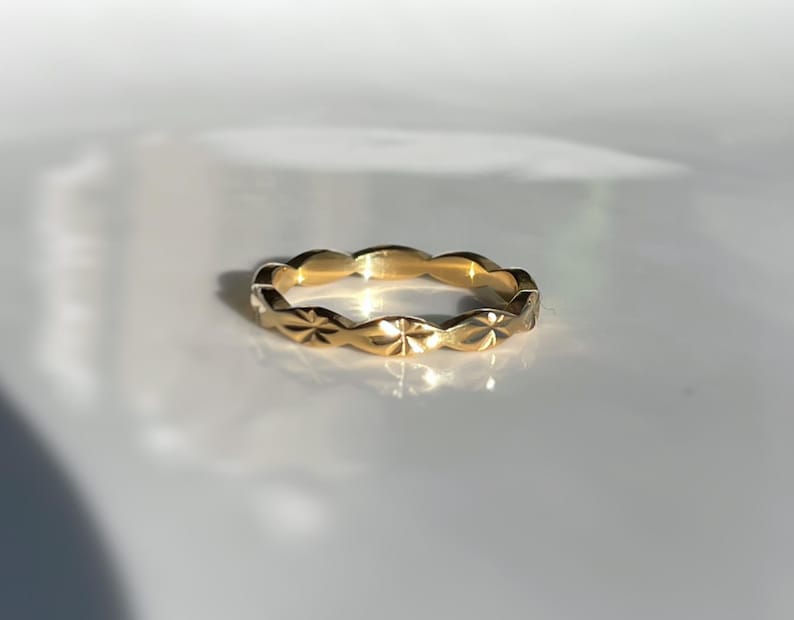 Peut inclure: Une bague en or avec un motif r&eacute;p&eacute;titif de bords festonn&eacute;s. Chaque feston est orn&eacute; d'une petite gravure en forme d'&eacute;toile qui capte la lumi&egrave;re, cr&eacute;ant une surface textur&eacute;e et r&eacute;fl&eacute;chissante sur toute la circonf&eacute;rence de l'anneau.