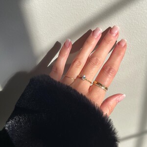 Peut inclure: Un ensemble de trois bagues dor&eacute;es port&eacute;es aux doigts. Une bague pr&eacute;sente une petite pierre claire en forme de poire, une autre est un anneau dor&eacute; fin et simple, et la troisi&egrave;me est un anneau dor&eacute; textur&eacute; avec une rang&eacute;e de petites pierres claires.