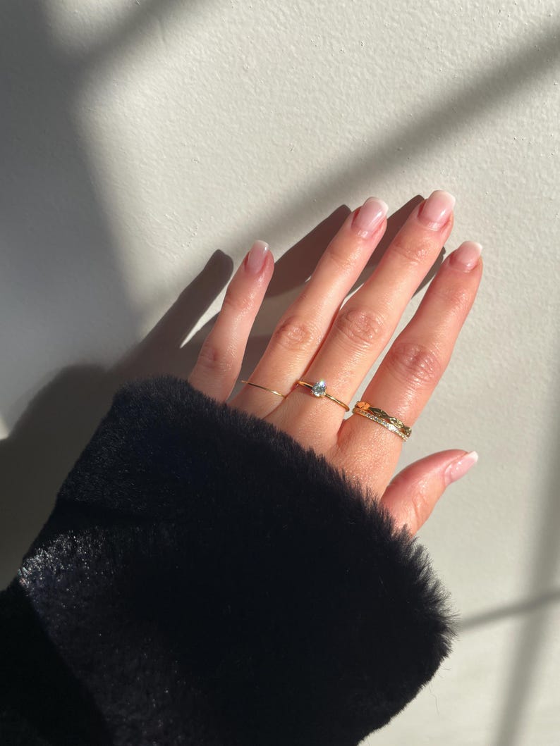 Peut inclure: Un ensemble de trois bagues dor&eacute;es port&eacute;es aux doigts. Une bague pr&eacute;sente une petite pierre claire en forme de poire, une autre est un anneau dor&eacute; fin et simple, et la troisi&egrave;me est un anneau dor&eacute; textur&eacute; avec une rang&eacute;e de petites pierres claires.