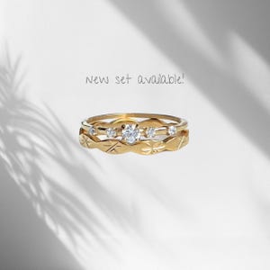 Peut inclure: Un ensemble de deux bagues en or. La bague sup&eacute;rieure pr&eacute;sente une pierre ronde transparente centrale entour&eacute;e de quatre pierres plus petites. La bague inf&eacute;rieure arbore un motif d'&eacute;toiles grav&eacute;es. Le texte New set available! est visible.