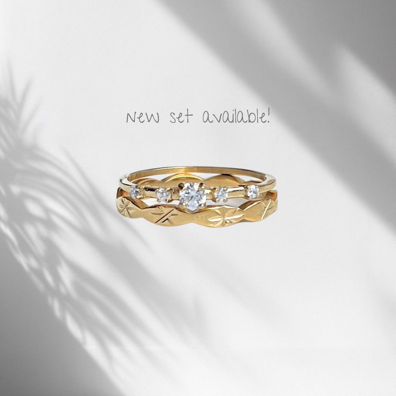 Peut inclure: Un ensemble de deux bagues en or. La bague sup&eacute;rieure pr&eacute;sente une pierre ronde transparente centrale entour&eacute;e de quatre pierres plus petites. La bague inf&eacute;rieure arbore un motif d'&eacute;toiles grav&eacute;es. Le texte New set available! est visible.