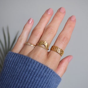 May include: Three gold-toned rings worn on fingers. One ring features a thin band with small clear stones, another is a sunburst design stacked with a plain band, and the third is a wide band with an intricate, open-work lattice pattern.