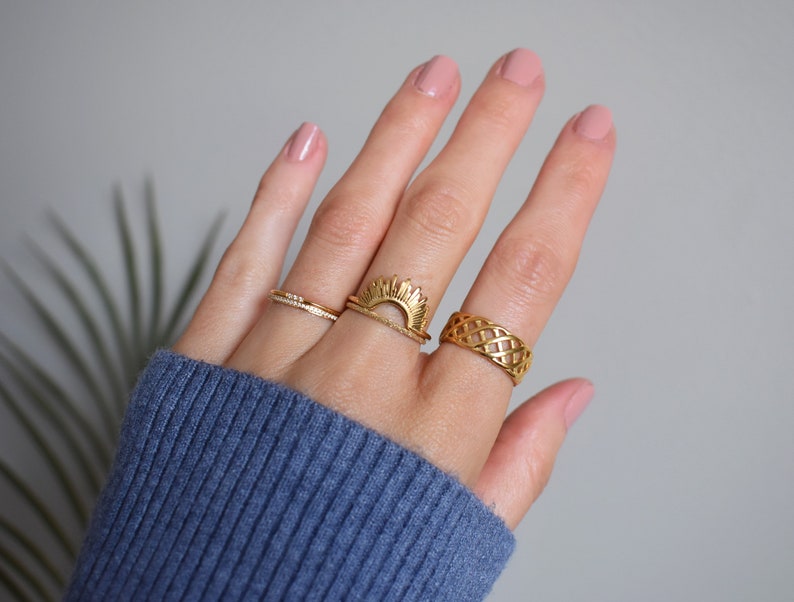 May include: Three gold-toned rings worn on fingers. One ring features a thin band with small clear stones, another is a sunburst design stacked with a plain band, and the third is a wide band with an intricate, open-work lattice pattern.