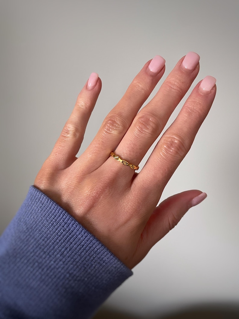 Peut inclure: Une fine bague en or pr&eacute;sentant un motif r&eacute;p&eacute;titif de petites &eacute;toiles ou de rayons de soleil grav&eacute;s sur toute la surface ext&eacute;rieure. Le m&eacute;tal a une finition polie et le design se compose d'&eacute;toiles alternant avec de petites indentations circulaires.