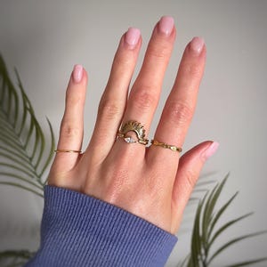 Peut inclure: Un ensemble de trois bagues dor&eacute;es port&eacute;es sur les doigts. La bague centrale pr&eacute;sente un motif en rayons de soleil avec une petite pierre claire taille marquise. L'index porte un anneau fin et l'annulaire une bague au motif textur&eacute;.