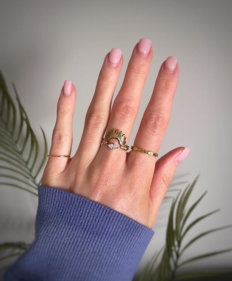 Peut inclure: Un ensemble de trois bagues dor&eacute;es port&eacute;es sur les doigts. La bague centrale pr&eacute;sente un motif en rayons de soleil avec une petite pierre claire taille marquise. L'index porte un anneau fin et l'annulaire une bague au motif textur&eacute;.