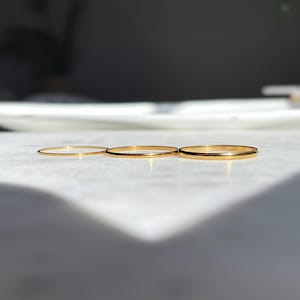 May include: Three thin, polished gold-tone metal rings arranged in a horizontal row. Each ring features a smooth, circular band with a reflective surface that catches the light, showcasing a simple, minimalist design with no additional embellishments.