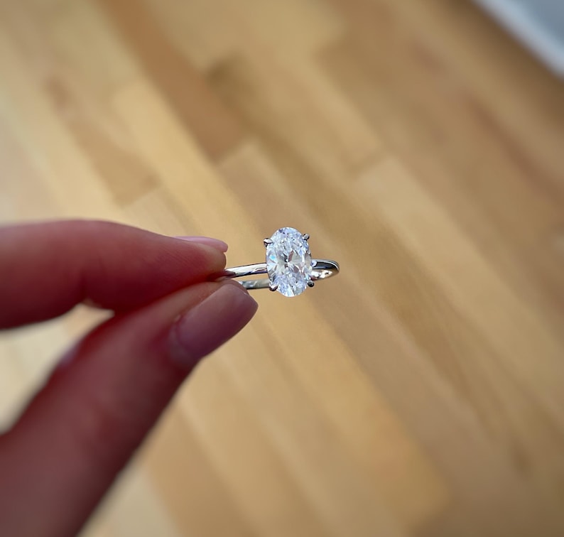 K&ouml;nnte beinhalten: Ein silberfarbener Ring mit einem einzelnen, klaren, oval geschliffenen Edelstein in einer Vier-Krappen-Fassung. Die Ringschiene ist schmal und poliert und weist eine reflektierende metallische Oberfl&auml;che auf, die den facettierten Stein betont.