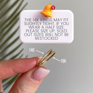May include: Two thin gold rings, one labeled 14K with a textured surface and one labeled 18K with a smooth surface. A white rectangular sign above reads: THE 14K RINGS MAY FIT SLIGHTLY TIGHT. IF YOU WEAR A HALF SIZE, PLEASE SIZE UP. SOLD OUT SIZES WILL NOT BE RESTOCKED.
