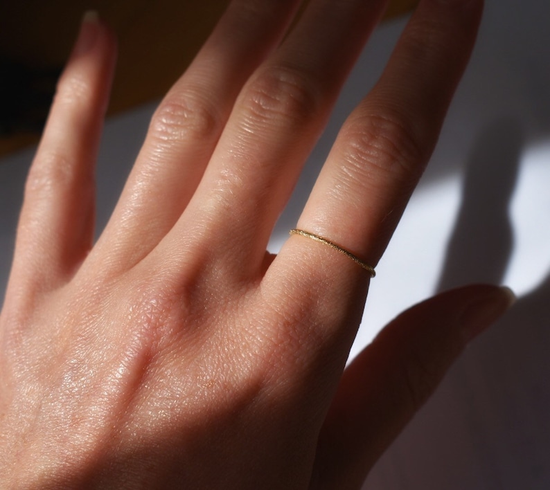 May include: A thin, gold-colored metal ring with a textured, sparkling surface finish. The band is narrow and circular, designed to be worn on a finger. The metal reflects light, creating a bright, metallic appearance across the entire ring.