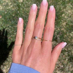 May include: A set of three gold rings worn on the fingers. One ring features a single round clear stone, another is a thin band with small embedded stones, and the third is a plain, very thin gold wire band. The rings have a polished finish.