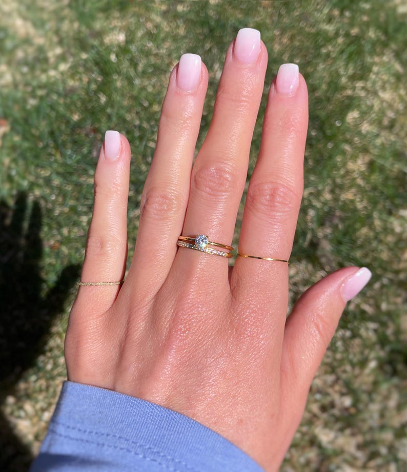 May include: A set of three gold rings worn on the fingers. One ring features a single round clear stone, another is a thin band with small embedded stones, and the third is a plain, very thin gold wire band. The rings have a polished finish.
