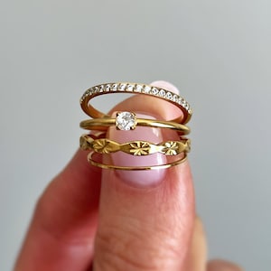 May include: A stack of four gold-toned rings. The top ring features a row of small clear stones. The second ring has a single round clear stone. The third ring displays a repeating floral or star-like pattern. The bottom ring is a plain band.