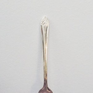 May include: A silver fork with a decorative handle, resting on a purple and black patterned textile. The fork has four tines and a detailed design at the top of the handle. The background is a light gray color.