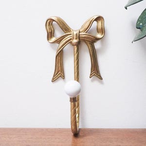 May include: A gold-colored metal wall hook with a bow design at the top and a white, round knob at the hook's end. The hook is mounted on a wooden surface against a white wall, with a green plant visible in the background.