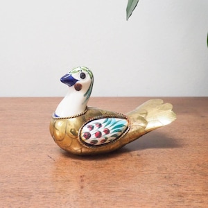 May include: A small, gold-coloured bird figurine with a white head and blue eyes. The bird is decorated with red, green, and blue floral patterns. The figurine is sitting on a wooden surface.