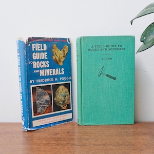 May include: Two vintage field guide books on rocks and minerals. One book is blue with the title "A Field Guide to Rocks and Minerals" and the other is green with the same title. The books are on a wooden surface.