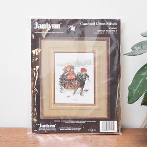 May include: A Janlynn Corporation counted cross stitch kit, featuring a winter scene of children with a sled. The packaging reads "Winter Reverie" and "Counted Cross Stitch". The frame is brown with a gold border. The kit is in a plastic sleeve.