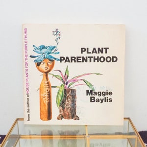 May include: A vintage book titled "Plant Parenthood" by Maggie Baylis. The book cover features an illustration of a potted plant and a wooden figurine with a blue flower on its head. The book is sitting on top of a glass display box with a gold frame.