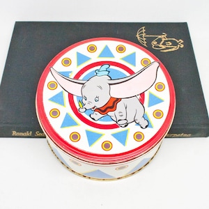 May include: A round tin featuring Dumbo the elephant from Disney. The tin has a red rim and a white base with a colourful pattern of blue triangles and yellow circles. The lid features a cartoon image of Dumbo.