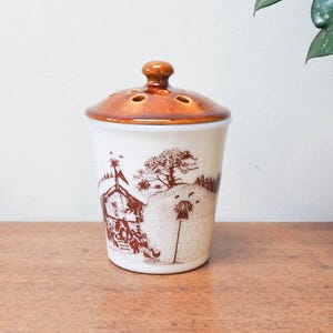 May include: A ceramic container with a lid, featuring a brown and white toile design. The container has a cylindrical shape with a brown lid and a small knob. The toile pattern depicts a rural scene with a house, trees, and figures. The container is on a wooden surface.