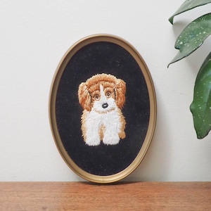 May include: Oval framed embroidery of a fluffy, seated puppy with brown and white fur, set against a black background. The frame is gold-toned. The artwork is displayed on a wooden surface.