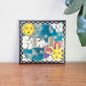 May include: A decorative light switch cover featuring a celestial design. The cover has a black border and a blue background with illustrations of a yellow sun, moon, clouds, and stars. The switch plate has two openings for the light switches.