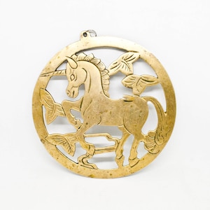 Puede incluir: Un adorno de metal de color dorado con forma de círculo con un diseño de unicornio recortado. El unicornio está de pie sobre sus patas traseras con la cabeza girada hacia un lado. El adorno tiene un lazo en la parte superior para colgar.