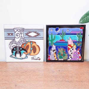 May include: Two decorative ceramic tiles. One tile features a Southwestern design with a vase, turquoise beads, and a gourd. The other tile depicts a desert scene with cacti, cats, and a night sky. The tiles are on a wooden surface.