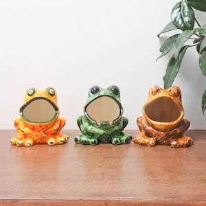 May include: Three ceramic frog-shaped planters in a row. The frogs are in shades of orange, green, and brown. Each frog has a wide open mouth and a hollow interior. The planters are on a wooden surface.