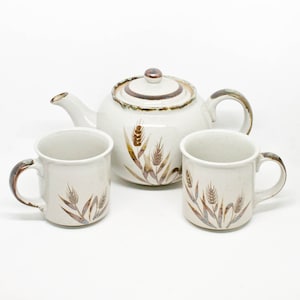 May include: A white teapot and two mugs with a brown wheat design. The teapot has a brown lid and a brown handle. The mugs have brown handles.