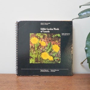May include: A spiral-bound book titled "Edible Garden Weeds of Canada" with a black cover featuring a photo of yellow dandelions. The book is on a wooden surface, with text from the National Museum of Natural Sciences.