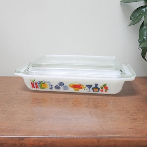 Könnte beinhalten: Eine rechteckige, weiße Pyrex-Auflaufform mit einem Deckel aus klarem Glas. Die Form hat ein farbenfrohes, vintage Design mit Obst und Gemüse. Die Form steht auf einer Holzoberfläche.