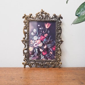May include: An antique brass picture frame with a floral print. The frame has detailed scrollwork and a dark background. The floral print features a bouquet of pink, white, and red flowers. The frame is displayed on a wooden surface.