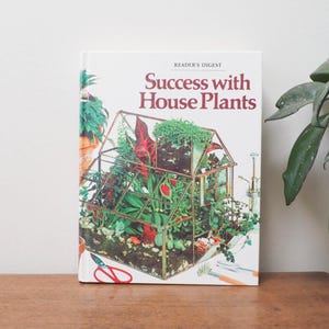 May include: A book titled "Success with House Plants" by Reader's Digest. The cover features a glass terrarium filled with various green and red plants. The book is on a wooden surface.