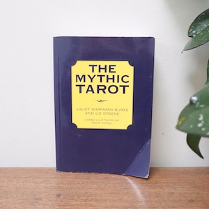 May include: A navy blue book titled "THE MYTHIC TAROT" with yellow text and a yellow rectangular border. The book is on a wooden surface. The author's names and illustrator are also listed on the cover.