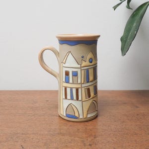 May include: A ceramic mug with a beige base and handle, featuring a hand-painted design of stylized buildings in shades of beige, blue, and brown. The rim is a light brown colour. The mug is on a wooden surface.