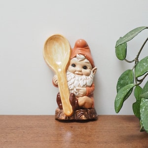 Puede incluir: Una figurita de gnomo de cerámica que sostiene una cuchara de madera. El gnomo lleva un gorro rojo y tiene barba blanca. El gnomo está sentado en un tronco marrón.