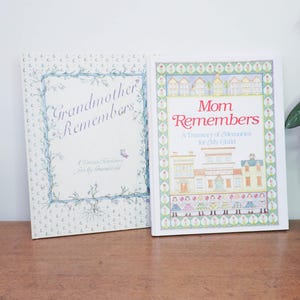 May include: Two memory books, "Grandmother Remembers" and "Mom Remembers," sit side-by-side on a wooden surface. The "Grandmother Remembers" book has a blue floral border and the text "Grandmother Remembers." The "Mom Remembers" book has a colorful illustration of houses.