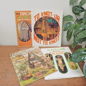 May include: A collection of vintage macrame books with colorful covers. Titles include "Macrame for All Seasons," "To Knot or Not to Knot," and "Macrame Magie." The books are arranged on a wooden surface, with a green plant in the background.
