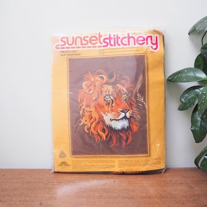 May include: A vintage Sunset Stitchery craft kit with a majestic lion design. The packaging is orange, brown, and yellow. The lion's face is in shades of orange, brown, and white. The kit includes the text "Majestic Lion" and "Lion Majestueux".