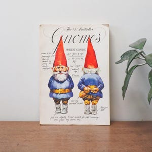 May include: A book cover with the title "Gnomes" in a whimsical font. The cover features two illustrations of gnomes with red hats, blue shirts, and brown belts. The text on the cover describes the gnomes as "Forest Gnomes" and provides details about their age and height.