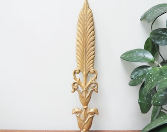 May include: A gold-colored, ornate wall decor piece in the shape of a stylized feather. The decorative element features intricate detailing, including a central vein and delicate curves. The piece is mounted on a white wall, with a wooden surface below.