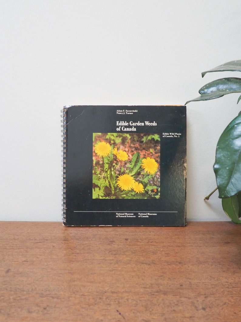 May include: A spiral-bound book titled "Edible Garden Weeds of Canada" with a black cover featuring a photo of yellow dandelions. The book is on a wooden surface, with text from the National Museum of Natural Sciences.