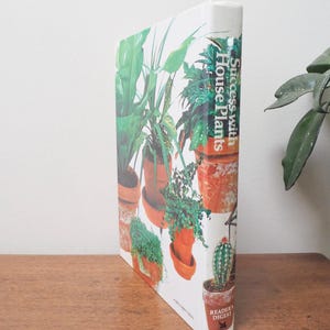 May include: A hardcover book titled "Success with House Plants" by Reader's Digest. The cover features a collage of potted plants in various sizes and shapes, with green foliage and terracotta pots. The book is on a wooden surface.
