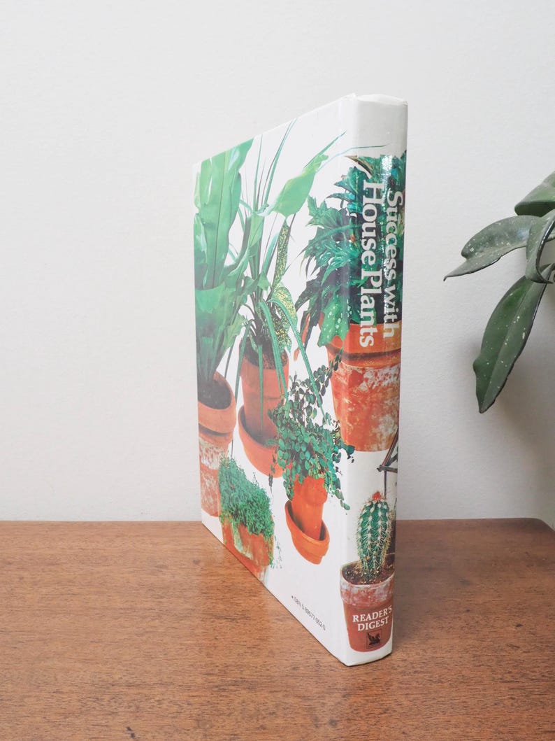 May include: A hardcover book titled "Success with House Plants" by Reader's Digest. The cover features a collage of potted plants in various sizes and shapes, with green foliage and terracotta pots. The book is on a wooden surface.
