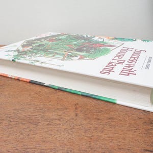 May include: A hardcover book titled "Success With House Plants" by Reader's Digest. The book's cover features a colorful illustration of a greenhouse with various plants. The book is resting on a wooden surface.